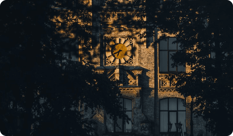 A clock on an old brick university building lit by the golden rays of the evening sun, seen through dark foliage.
