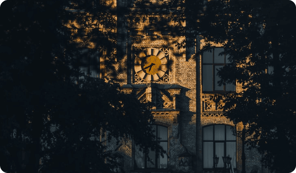 Годинник на старовинній цегляній будівлі університету, що освітлений золотими променями вечірнього сонця, вид через темне листя дерев.